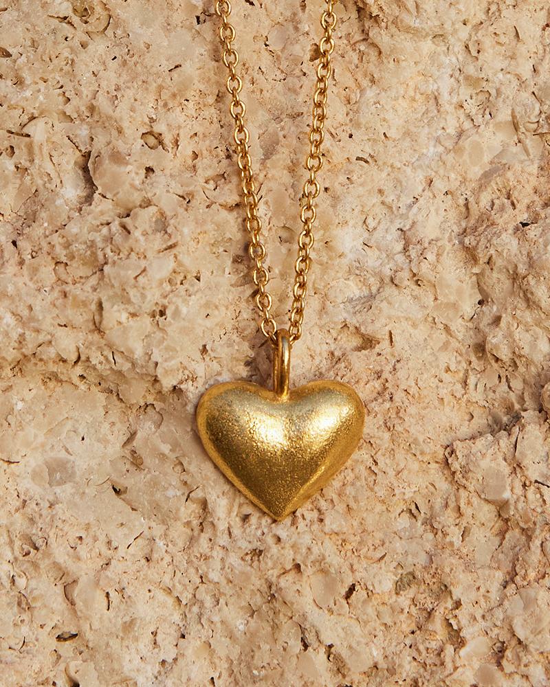 Leona Necklace: Gold chain with a matte, textured gold heart pendant on a light stone background.