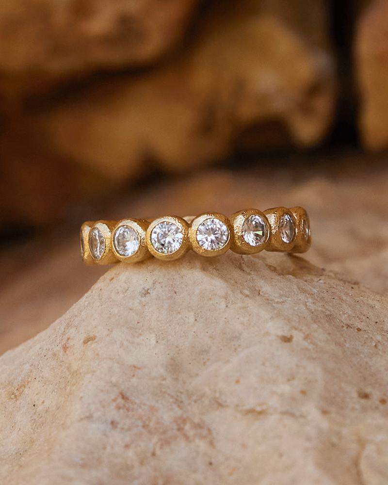 Anello Amélia con una fila di pietre rotonde scintillanti incastonate a castone in una fascia dorata testurizzata, posato su una superficie in pietra.