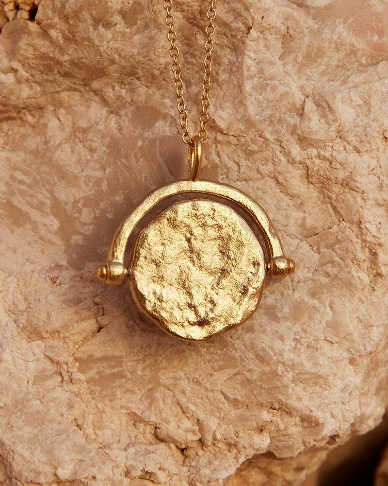Gold Solune necklace with a textured, round rotating pendant on a delicate chain, against a light brown rock.