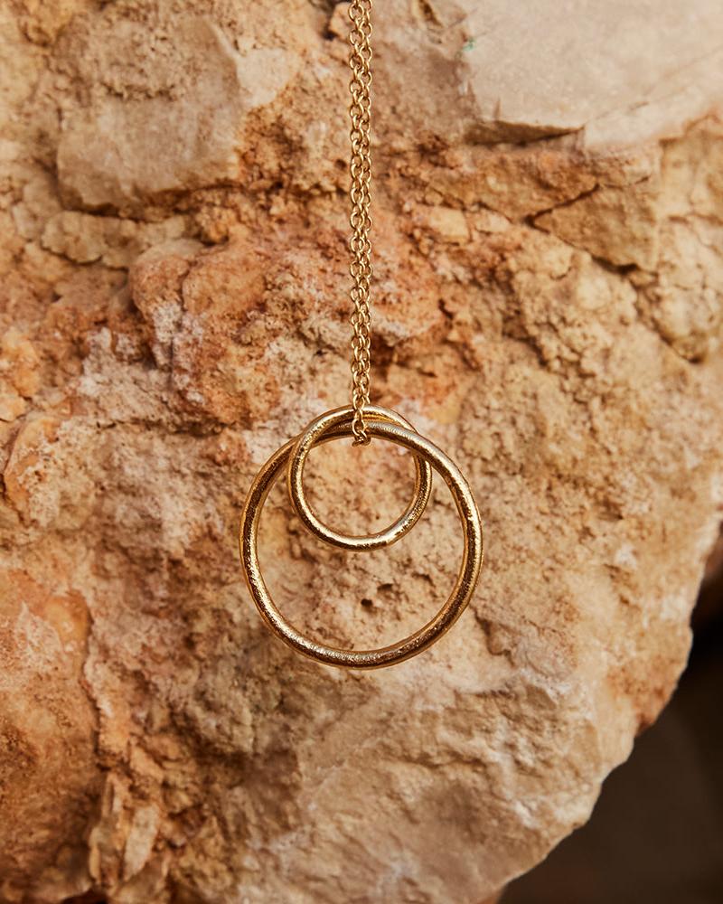 Selina gold necklace with two interlocking textured rings on a natural rock background.