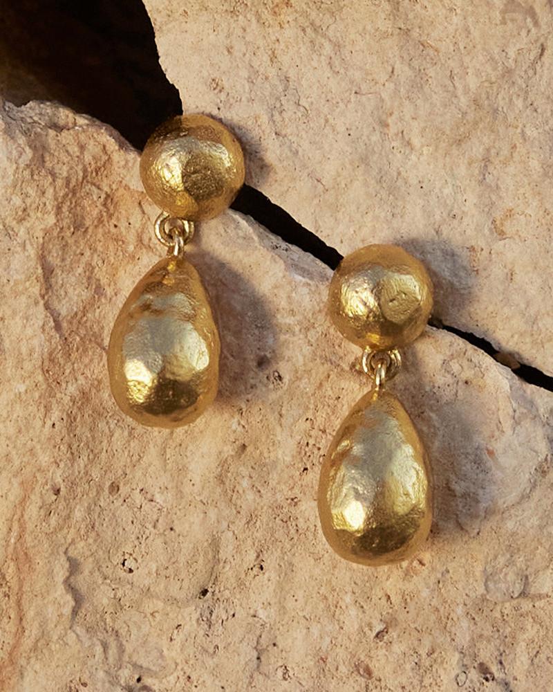 Dalias gold earrings with textured spherical studs and organic teardrop pendants on a stone background.