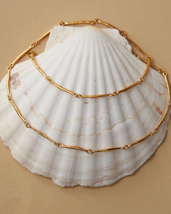 Enea gold-tone necklace with slim, hammered bar-shaped links, displayed on a white seashell.