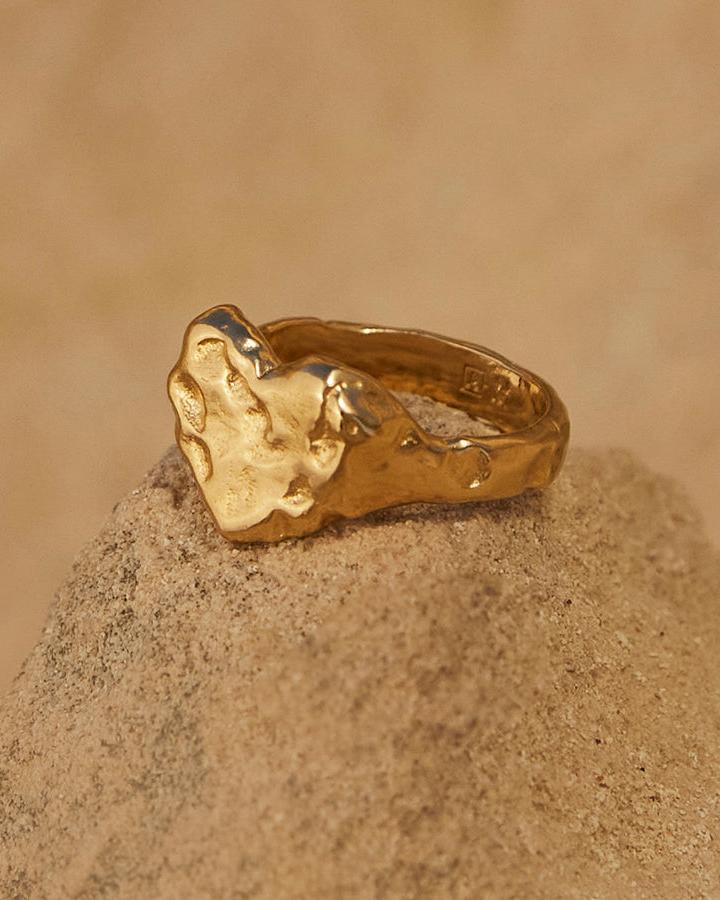 Chunky gold Livia heart signet ring with a sculptural, hammered finish on a sandy rock.