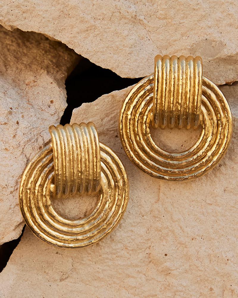 Giulia chunky gold dangling hoop earrings with ridged design, resting on light-colored rocks.