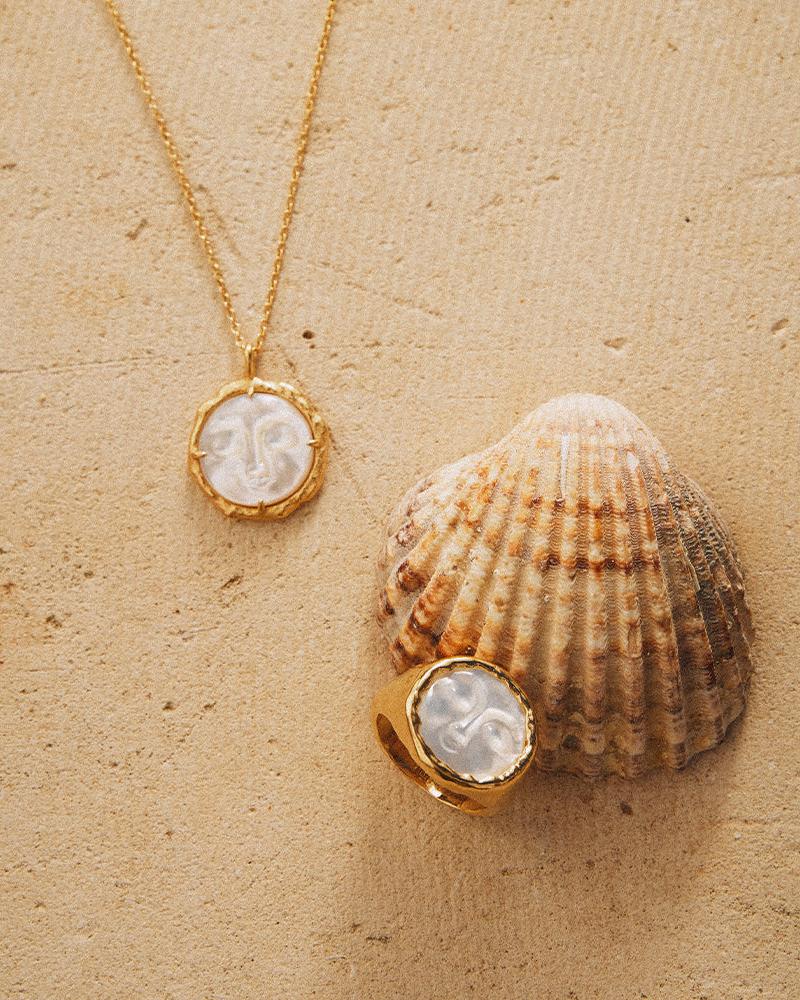 Golden Hëna ring with a carved mother-of-pearl moon face, next to a matching necklace and seashell on sand.