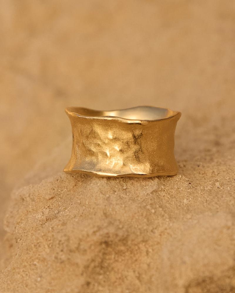 Gold Lucca ring, wide and concave with a hammered finish, on a sandy surface.