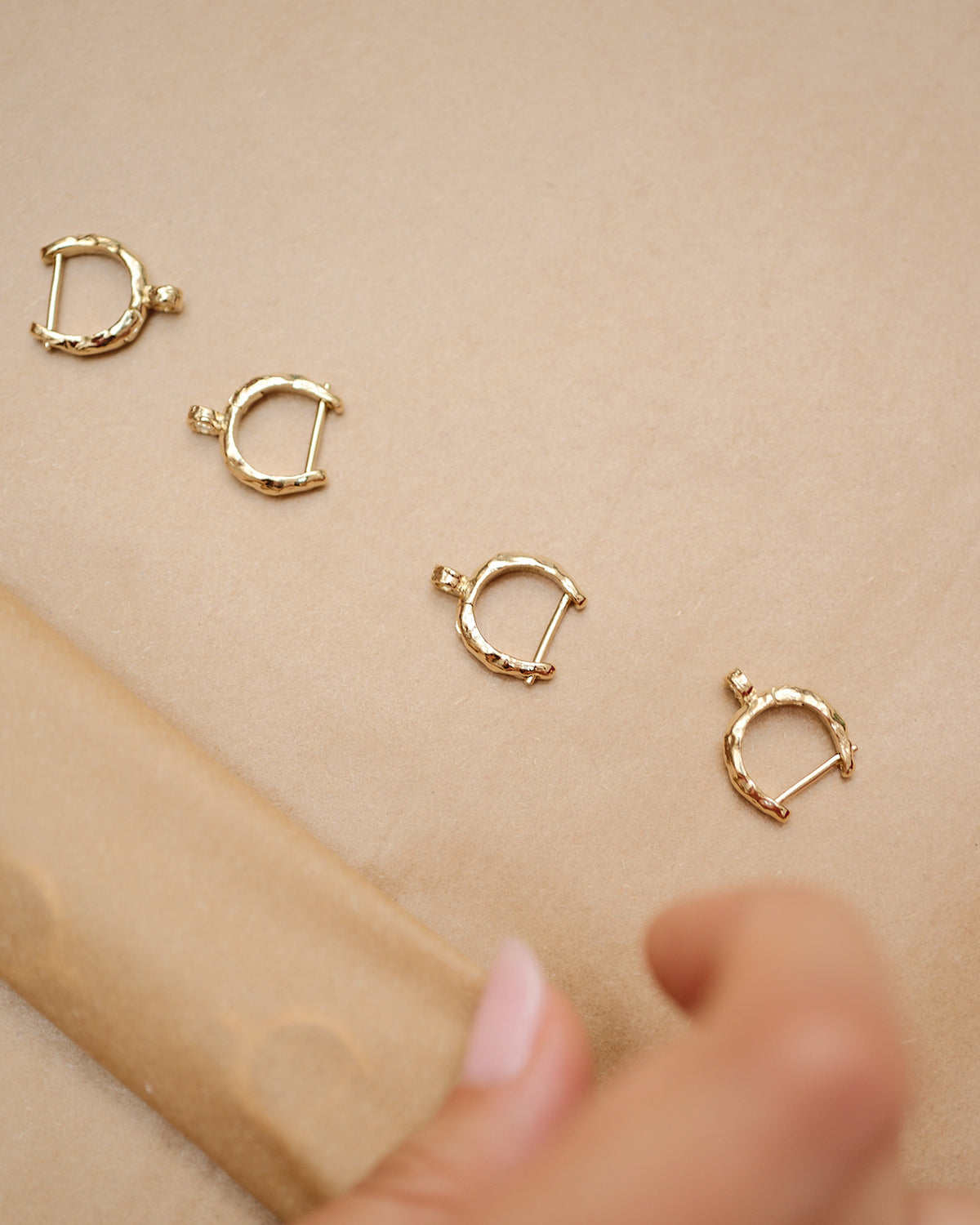 Four small, textured gold hoop earrings arranged diagonally on a neutral beige background with a hand in the foreground.