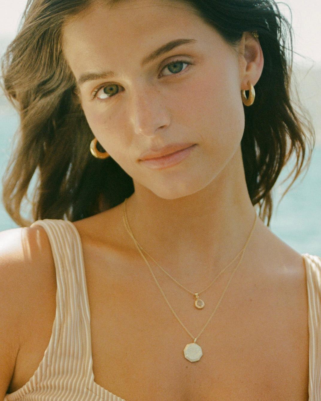 Woman wearing Aura open hoop earrings with a hammered gold finish and two layered gold necklaces.