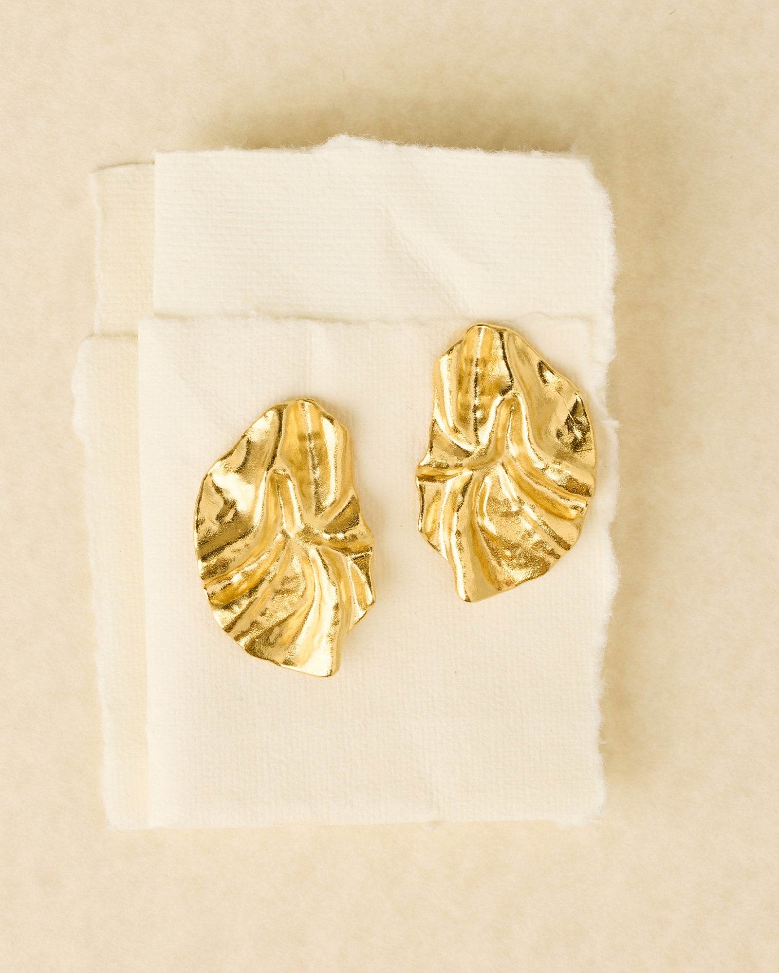 Gold Batla earrings with a soft petal shape and delicate fabric-like texture, displayed on cream paper.