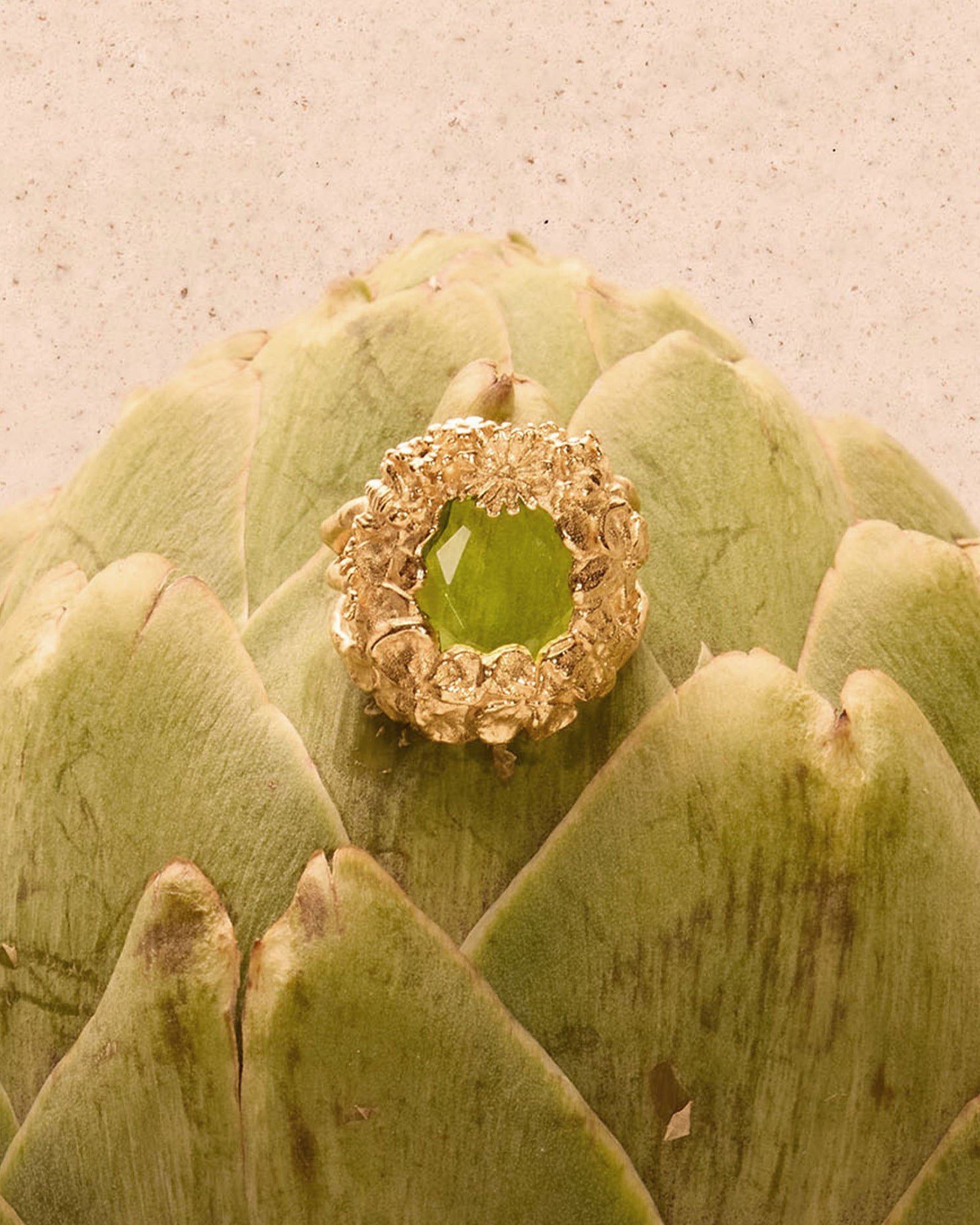 Hespé chunky gold ring with a striking green stone, nestled in a floral design, on an artichoke.