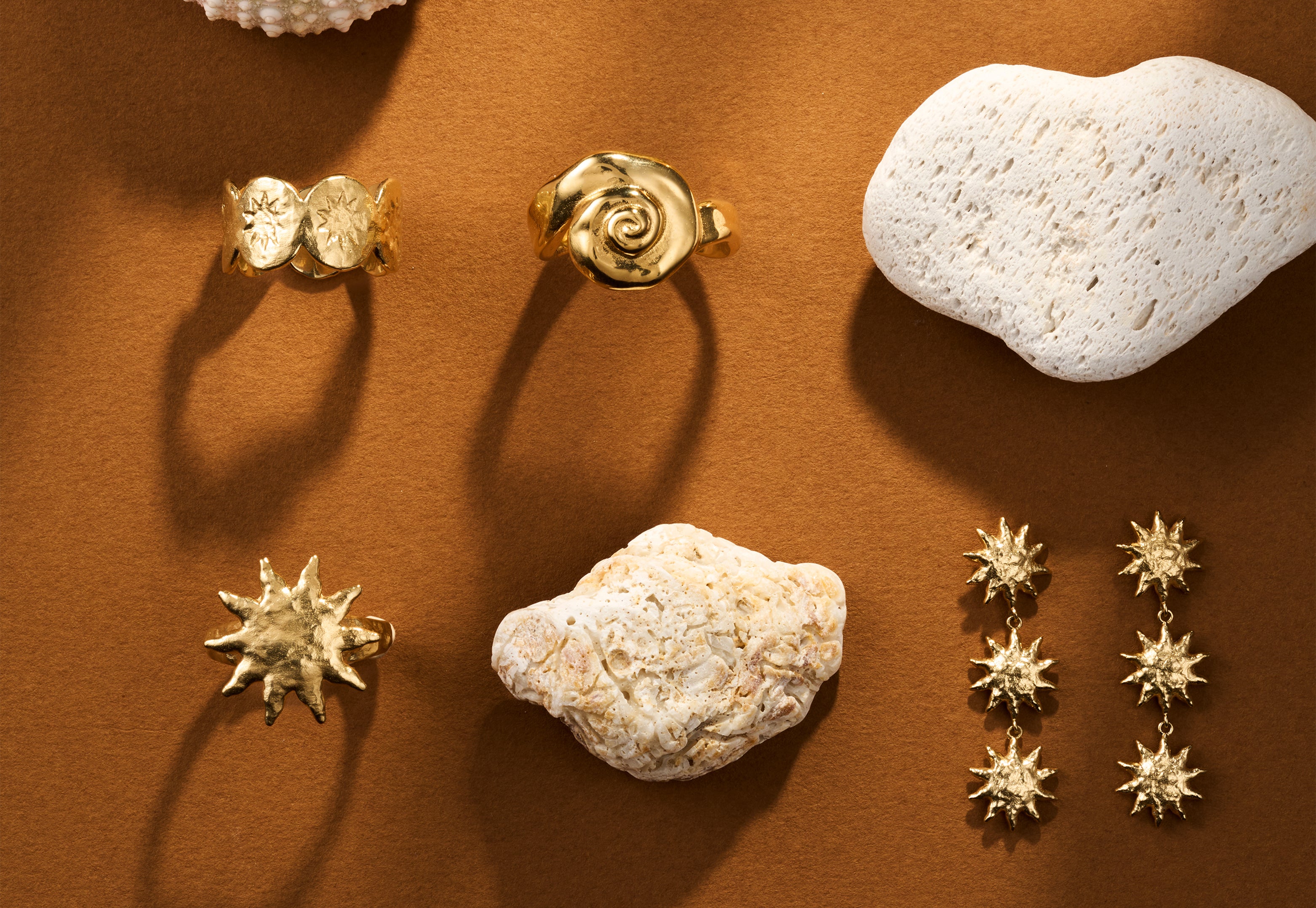 Gold jewelry featuring sun and spiral motifs arranged with textured white stones on a warm brown background.