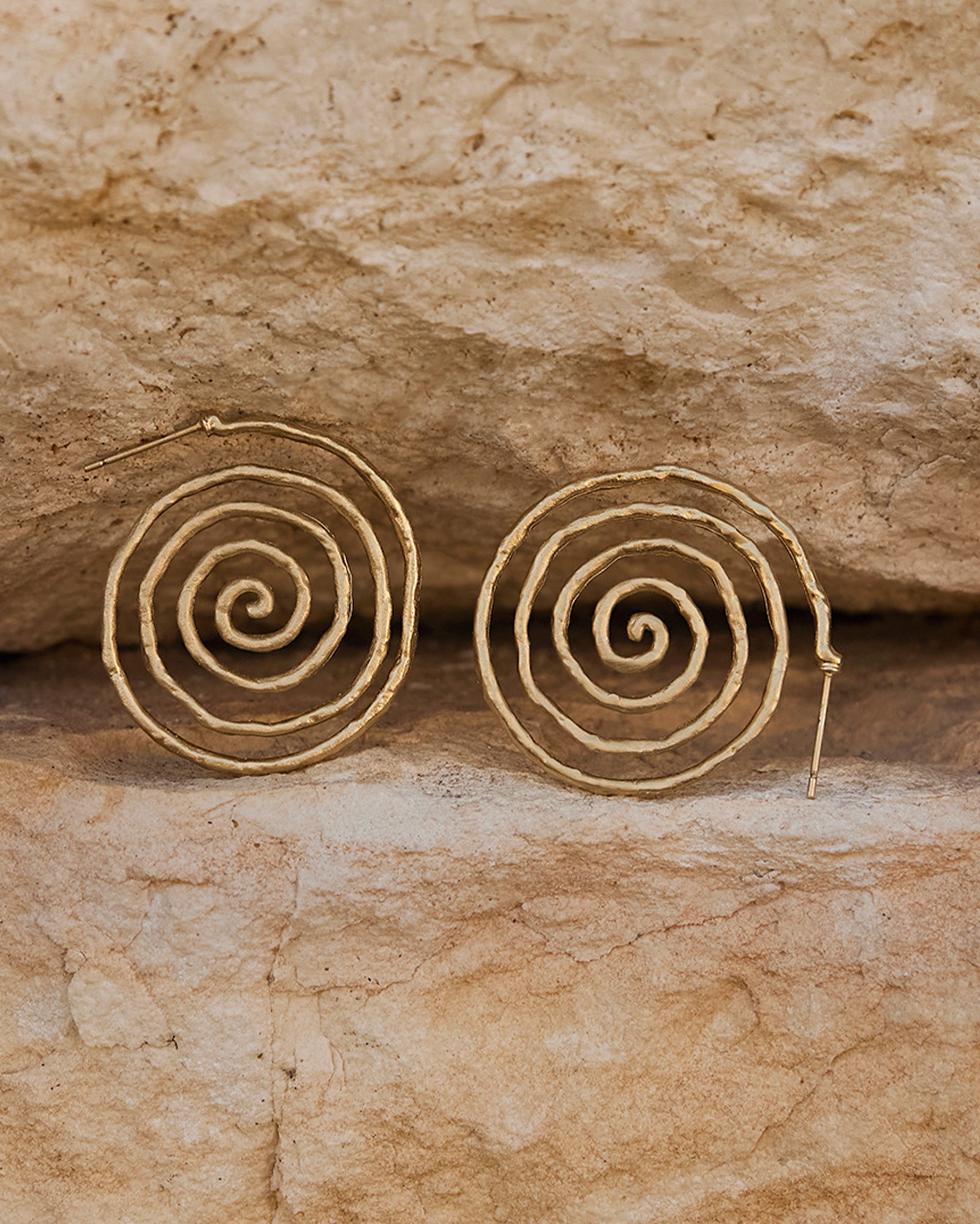Gold Nacci spiral earrings, fluid and curvy, on a textured light brown stone background.
