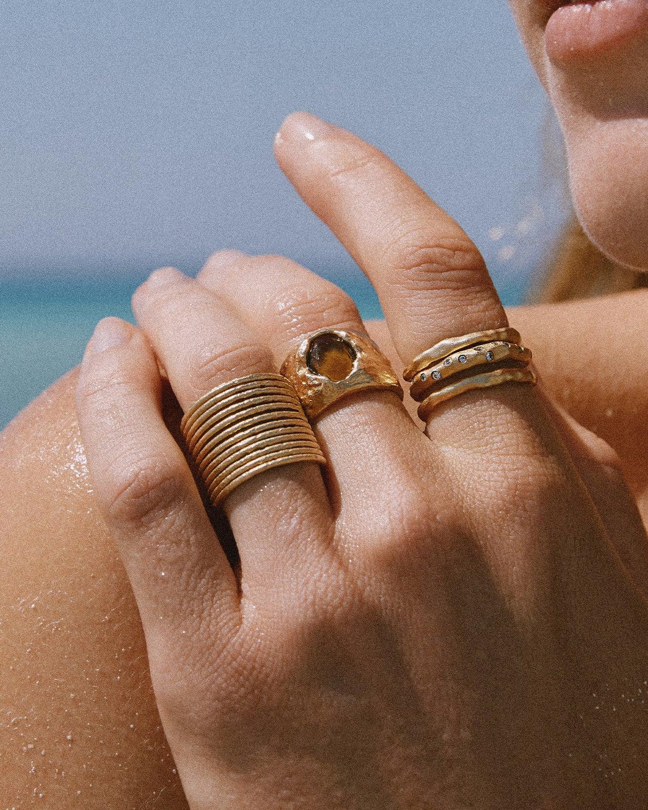 Verstellbarer, goldfarbener Etérea Ring mit 12 zarten, gestapelten Bändern, an einem Finger vor einem Strandhintergrund getragen.